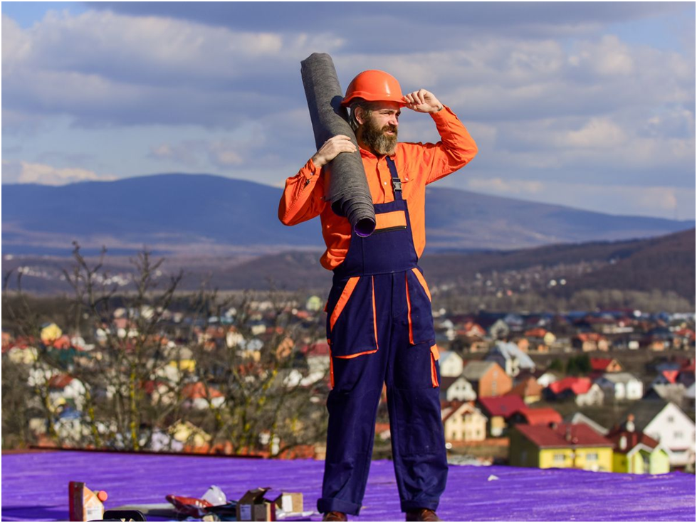 Roofer in safety gear carrying roofing roll on rooftop