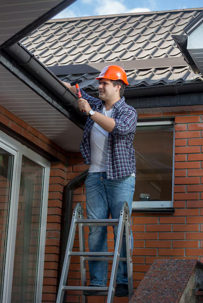 Roofer on ladder repairing roof gutter with tools